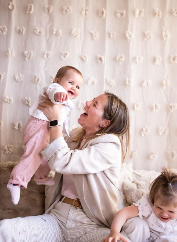 Mama tilt haar lachende baby op, met de grote zus ernaast