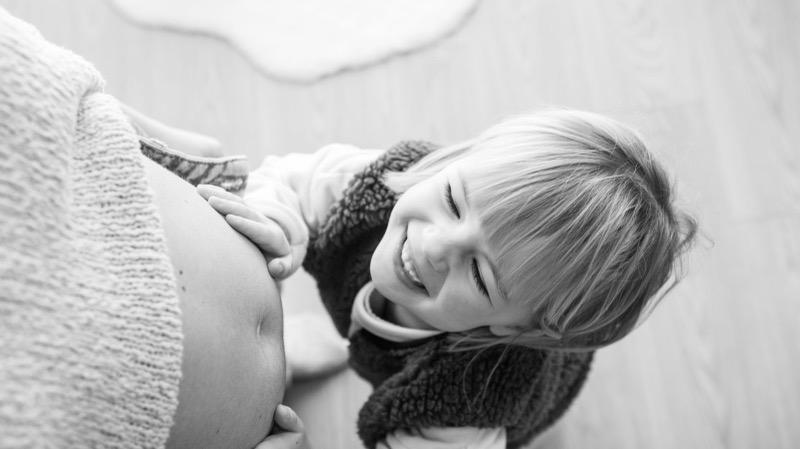 Zwart-witfoto van een lachend dochtertje naast de babybuik